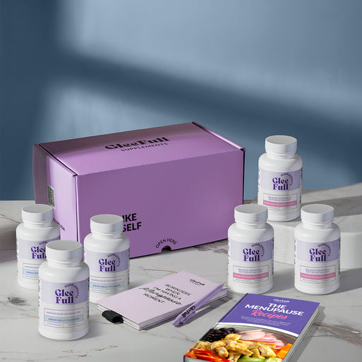 A product bundle display for GleeFull Supplements on a marble surface against a blue background. The layout includes a large purple shipping box featuring the slogan 'FEEL LIKE YOURSELF AGAIN.', three white bottles of Over 30 Hormone Support and three white bottles of Meno Belly Flusher, a purple branded pen, a matching notepad that reads 'REMINDERS WHEN I'M HAVING A MENOPAUSE MOMENT', and a recipe booklet titled 'THE MENOPAUSE Recipes'