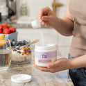 A person scoops white powder from a GleeFull Hormonelyte jar over a glass on a marble countertop, with a yogurt bowl, water glass and fresh strawberries beside it. Label reads 'Enhances Nutrient Absorption, Hormonelyte'.